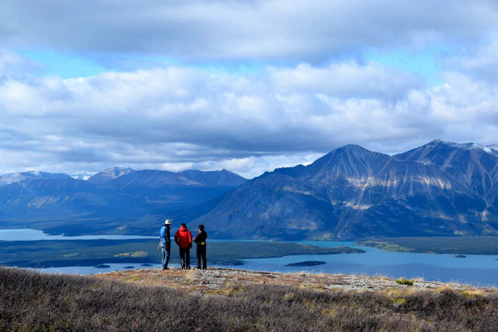 Photo: three scientists stand atop a bluff, overlooking a wide valley that separates them from a rugged mountain range