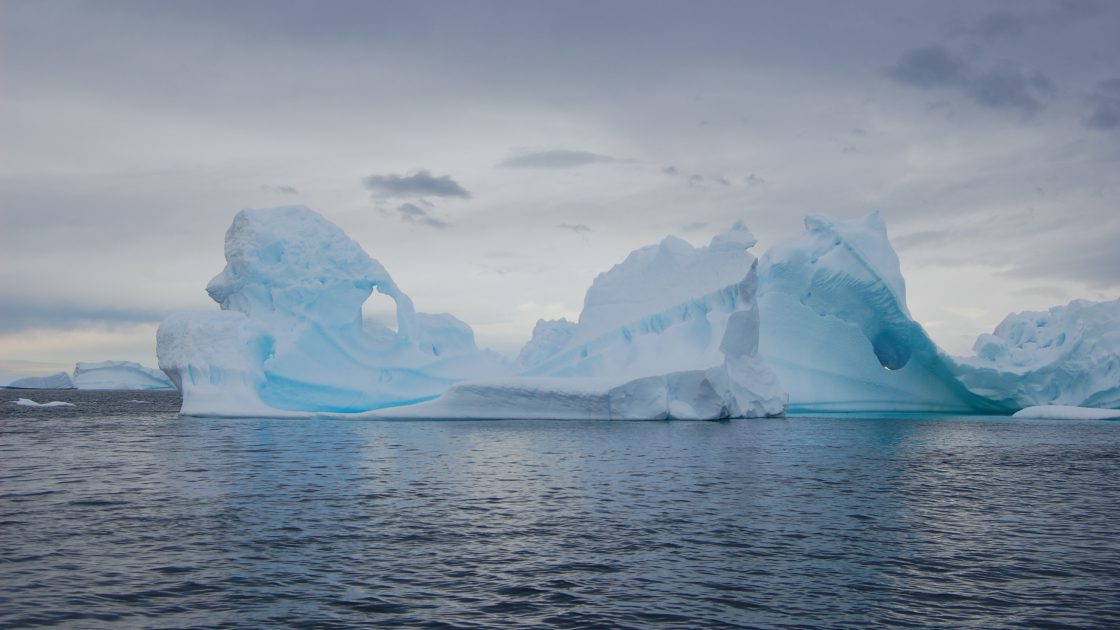 Photo: an old, weathered iceberg floats in a dusky sea, the iceberg carved by the elements to reveal delicate arches and rippling edges