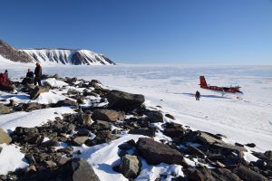 Photo: a group of scientists sit on a rocky outcrop overlooking a vast ice-locked ocean upon which a small research plane has landed