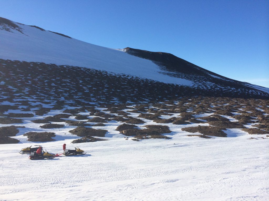 Photo: a scientist attends to three snowmobiles in front of an icy, low-slung hill
